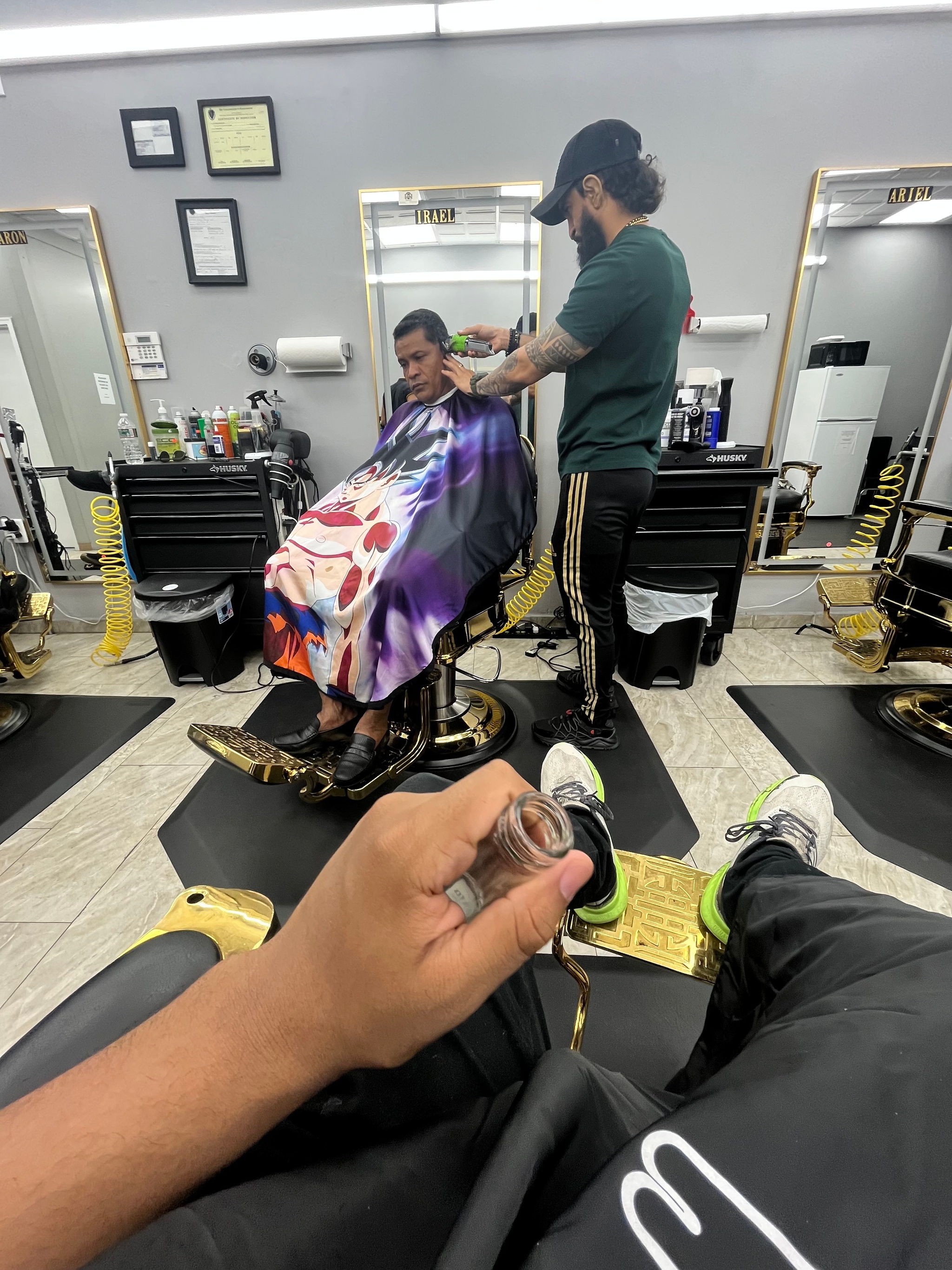 A scene in a barbershop taken by someone sitting in a chair across from a barber cutting the hair of a client. At the bottom of the image the photographer's hand protrudes into the image. The photographer, artist Bryan Fernandez, has light brown skin. 