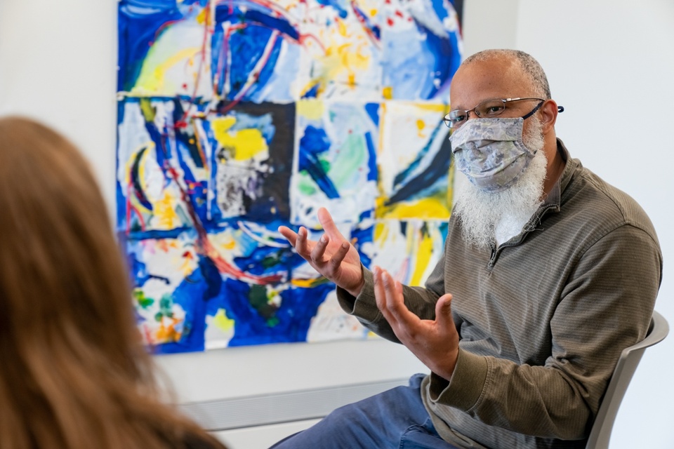 Instructor sits in front of a blue and yellow abstract painting hung on a white wall and speaks to a person just off camera.