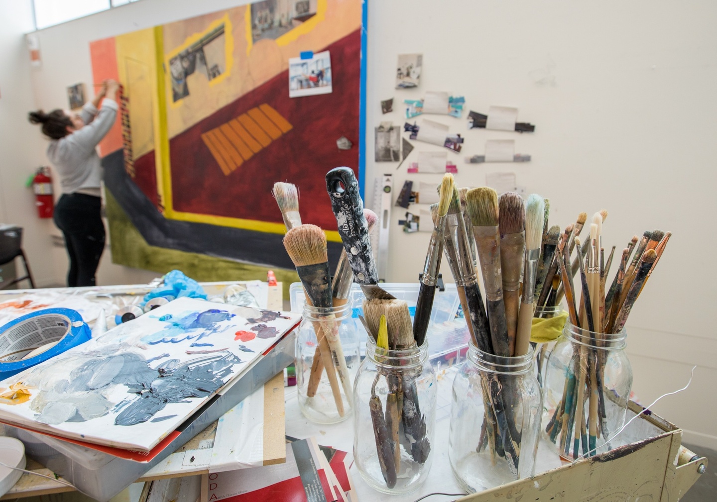 Artful shot of jars of brushes on a table next to a student working on a large-scale painting.