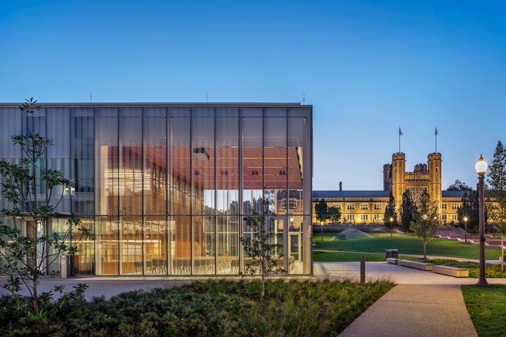 A 2-story glass-walled building seen in the foreground with the Collegiate Gothic Brookings Hall in the background, at sunset.