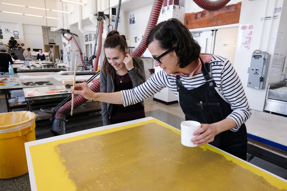 Person in black apron demonstrates dripping liquid onto a printmaking plate to another person.