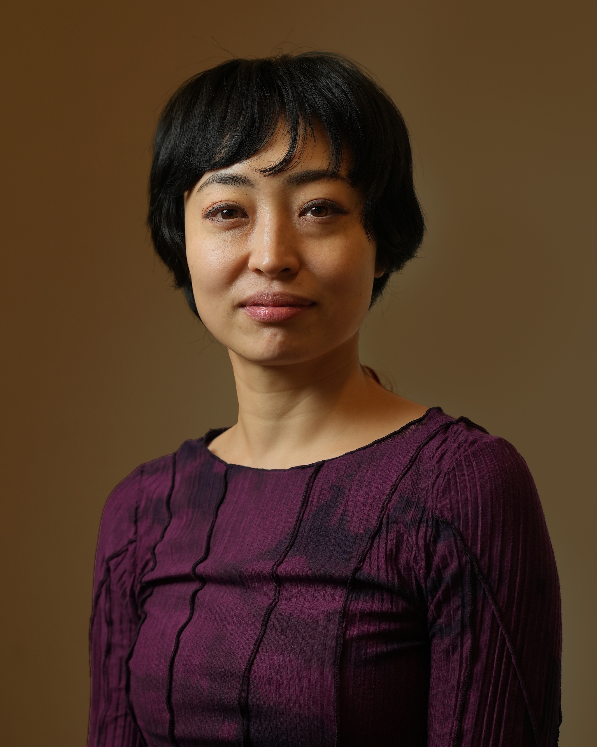 A portrait of Yukari Osaka against a neutral brown background. Yukari wears a dark purple shirt and looks directly at us with a closed-mouth smile. 