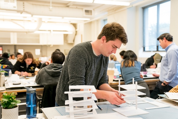 Student carefully assembles a model at a space in a large open format classroom filled with students.
