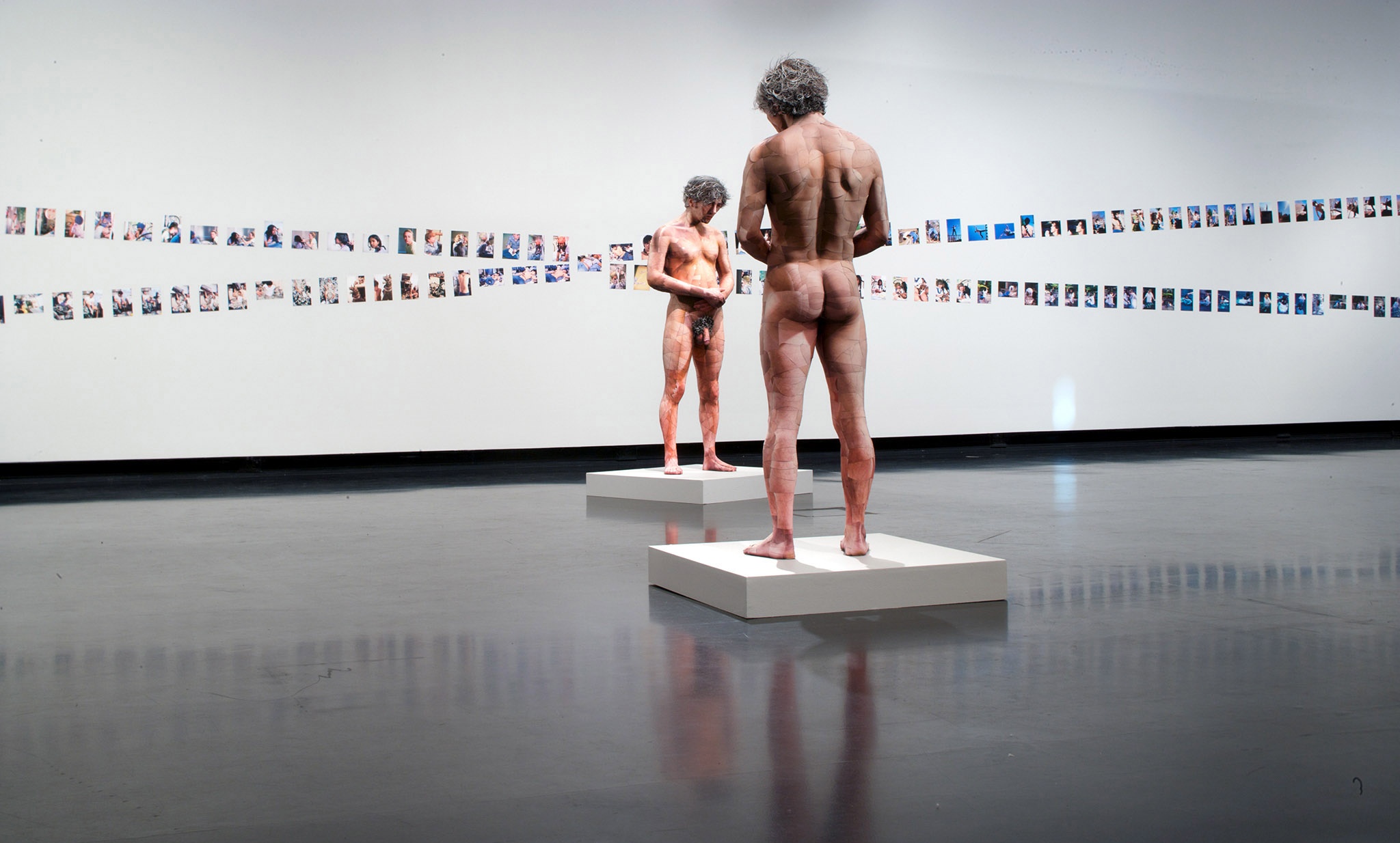Two life-sized, nude models made of flesh-colored photographs of a white man face each other in front of a white wall with two rows of photographs.