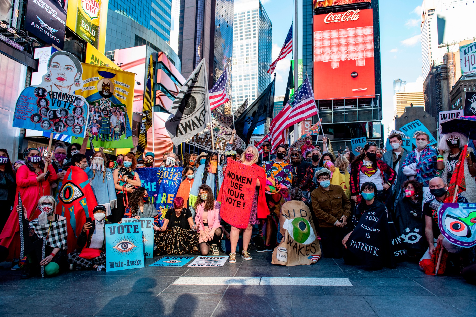 A photograph of a colorfully-dressed crowd in front of skyscrapers, holding flags and banners, including an American Flag, a “WIDE AWAKES.” banner, and a poster saying “WOMEN USE YOUR VOTE!”