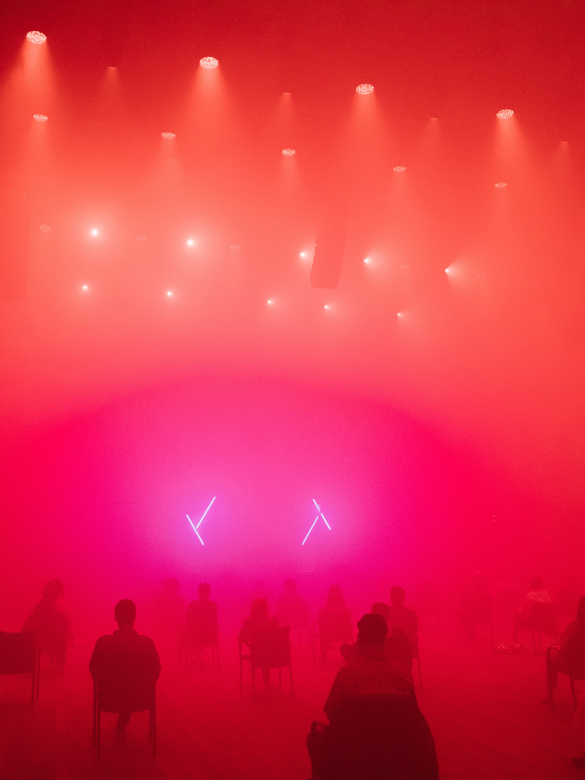 An audience seated in distanced seats in a foggy performance space lit with diffuse red, orange, and purple lighting