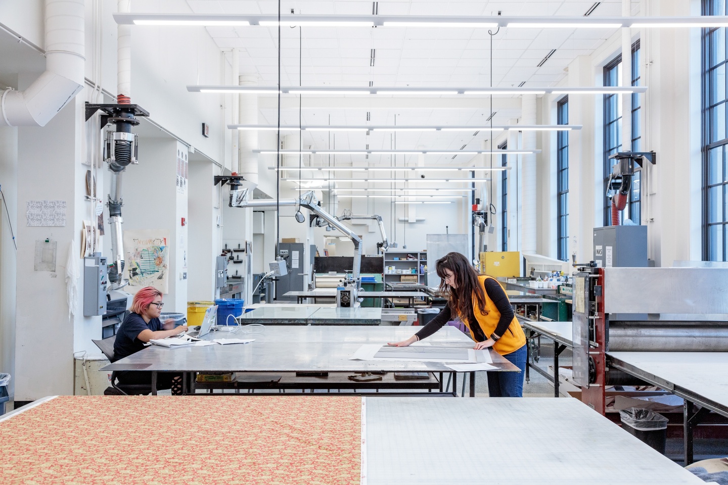 Printmaking studio space with white walls, tall ceilings, and lots of natural light. Two people look at prints on a work table.