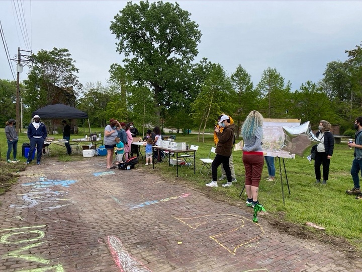 People gather around a pop-up tent with tables and displays on an empty lot with a strip of cobblestones. Children have made chalk drawings on the cobblestones. A few people inspect a posterboard with an architectural rendering on an easel.