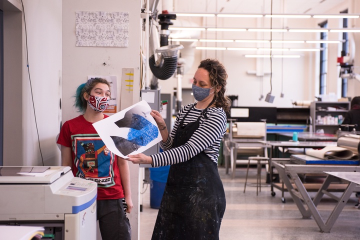 A person in a black apron stands next to a risograph machine with a person with blue hair and holds up a blue and black risograph print. Behind them is a large, well-lit space filled with printmaking presses and other equipment.
