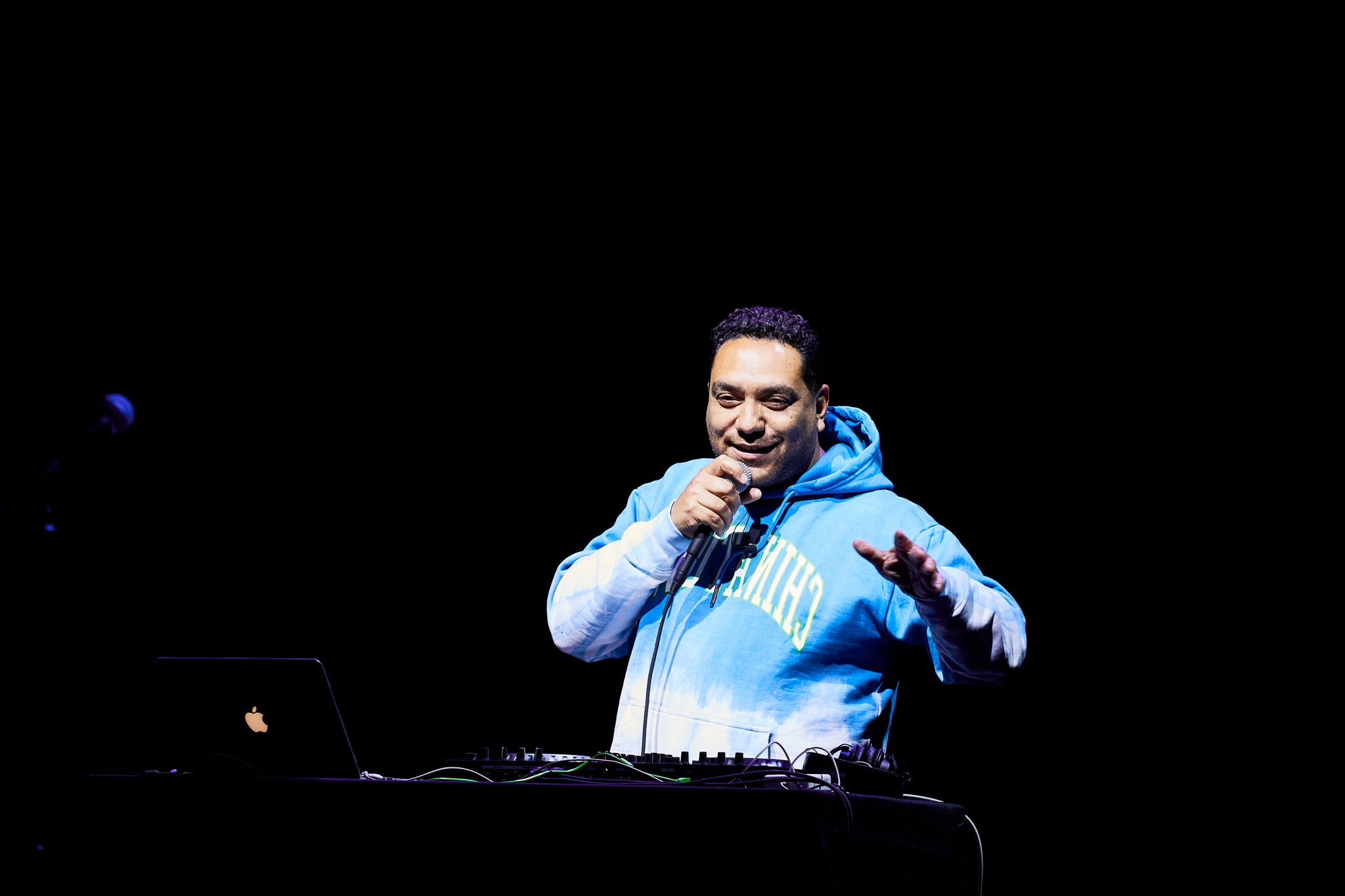 A DJ in a blue and white hoodie speaking into a microphone in front of his turntables