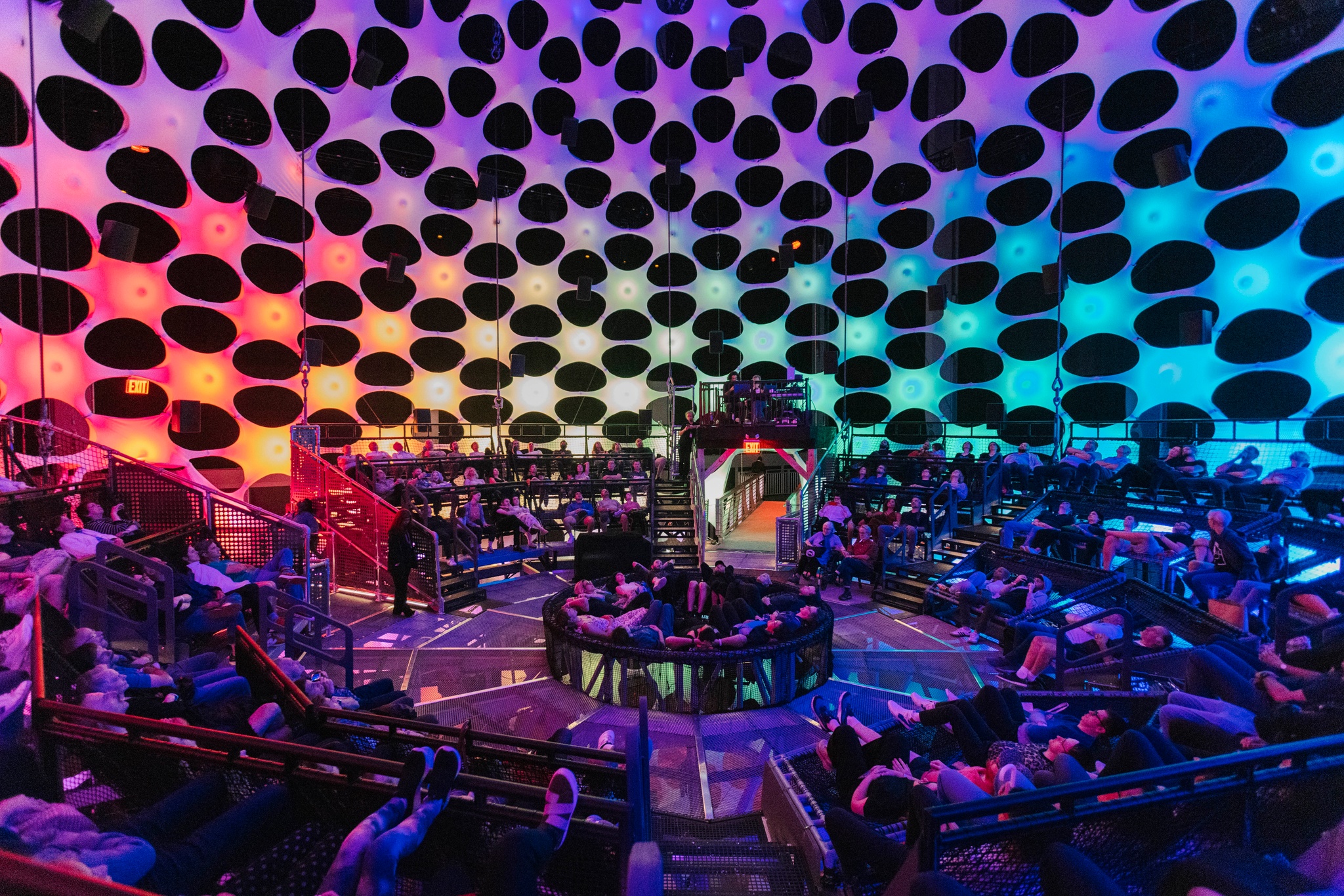 People in relaxed poses lie on their backs on netted seating banks inside a concert hall with a spherical wall. The curved wall towers above the audience and is porous and composed of light nodes that emanate orange, blue, and purple light.