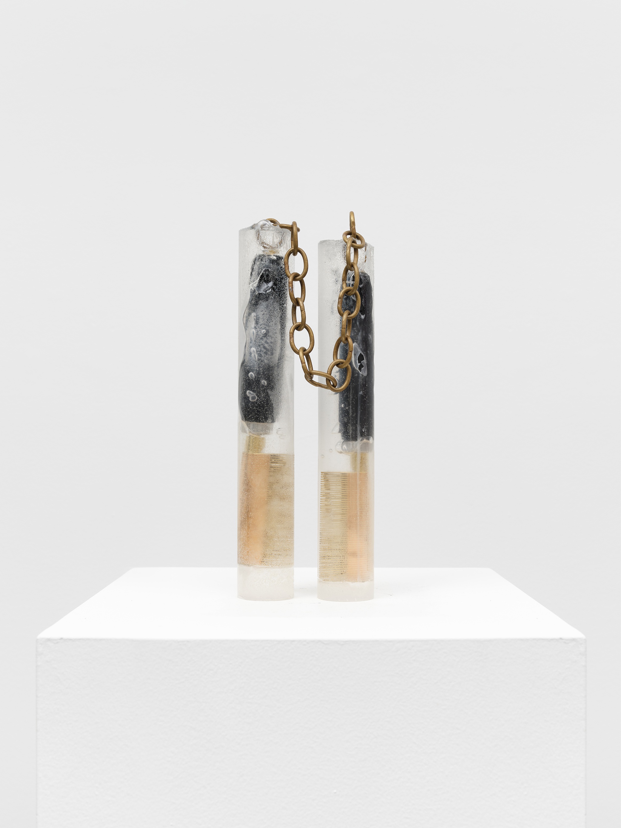A sculpture resembling nunchuks, with the bars in resin containing wood and brass hot combs. The two pieces are linked by a brass chain.