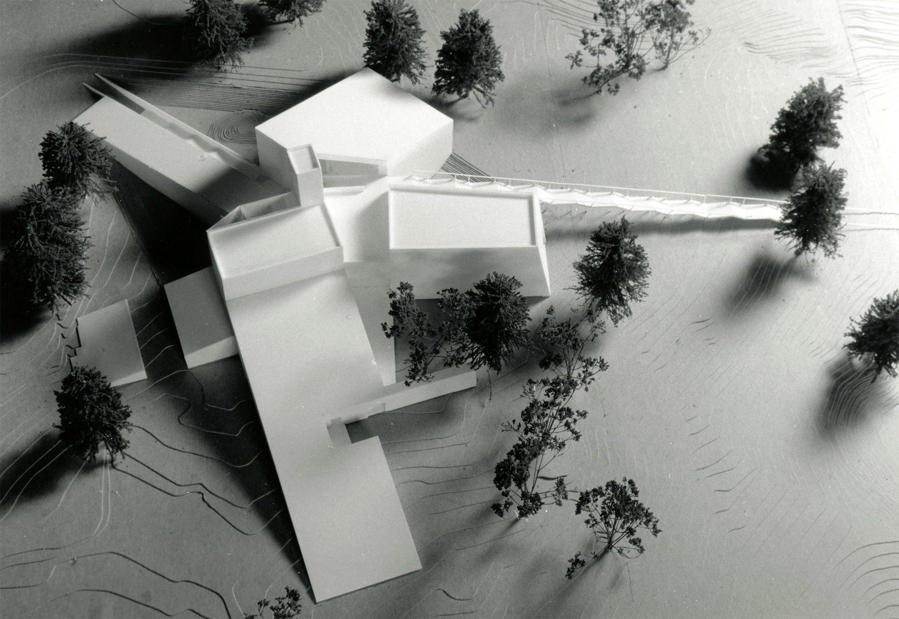 A black and white aerial photograph of a small, white, model of an angular building surrounded by fake trees.