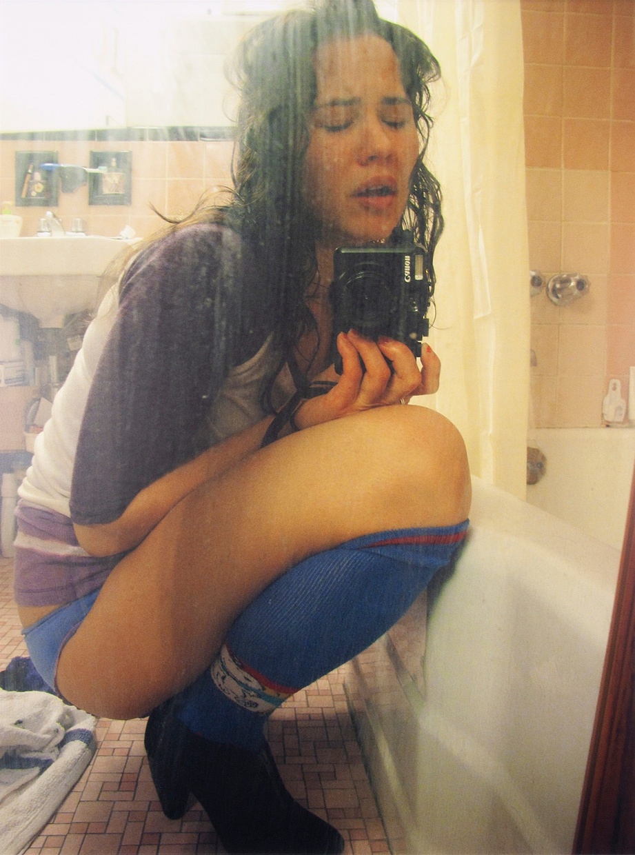 A woman crouches in a bathroom, crying while holding a camera.
