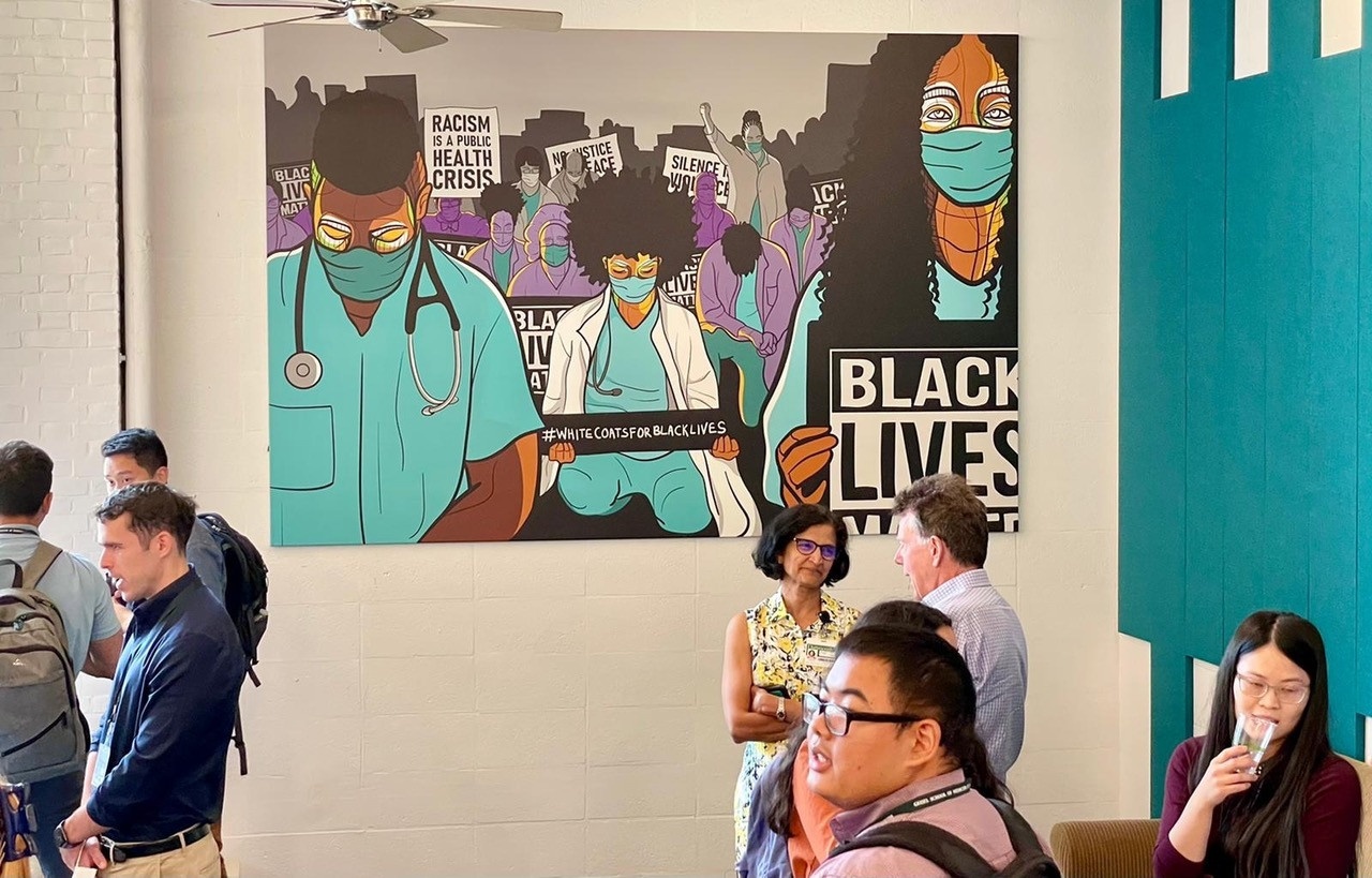 Installaed mural showing a collection of people in scrubs, white coats, and with other tools of the medical profession kneeling together. The people hold signs that read "#WhitecCoatsForBlackLives", "Racism is a Public Health Crisis," and "Black Lives Matter." People are seated at tables in front of the mural. 