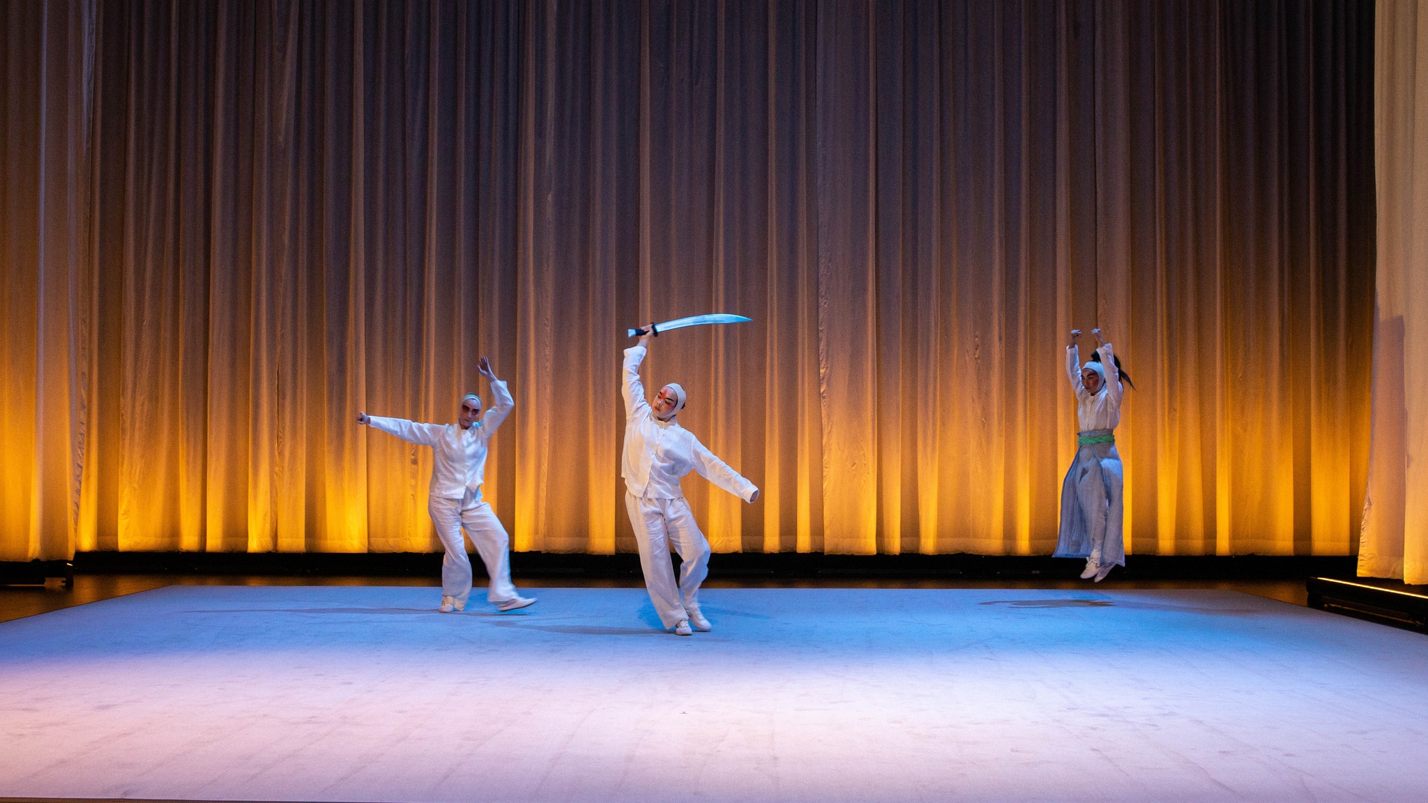 Three dancers in costumes inspired by Peking Opera appear on stage with a curtain lit with a golden light behind them. One stands with one leg outstretched, with one arm held parallel to the floor and one curved above her head. The Second dancer holds a sword above her head,, curving her body slightly. The third jumps straight up into the air with arms above her head.