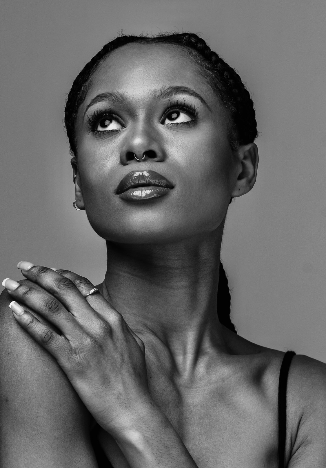 A woman with bare shoulders in a black tank top poses for a black-and-white headshot looking up with her right hand brought to rest on her left shoulder