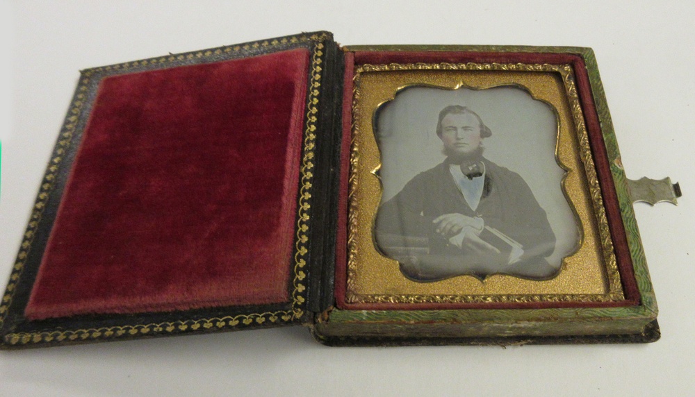 A red velveted gold case, opened like a book, holds a photograph of a man posing with one arm resting on a stack of books and another book in hand.