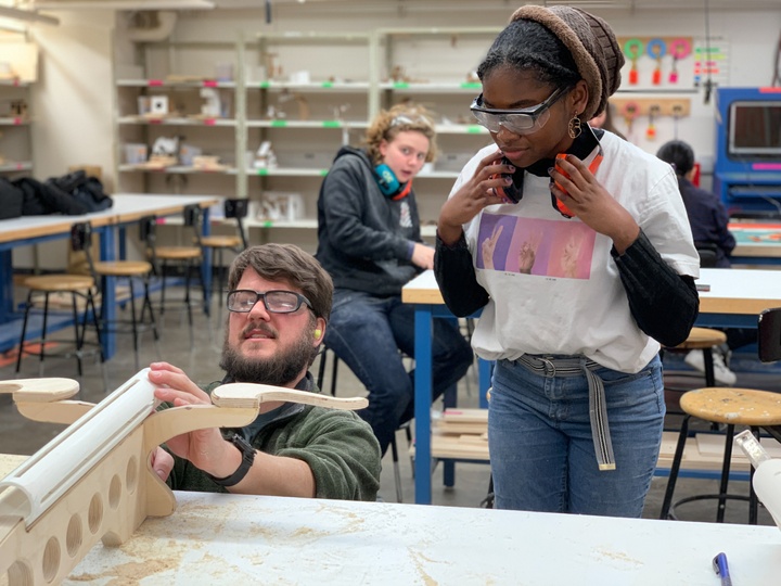 People with goggles and ear protection analyze an object made of PVC and wood in a woodshop.
