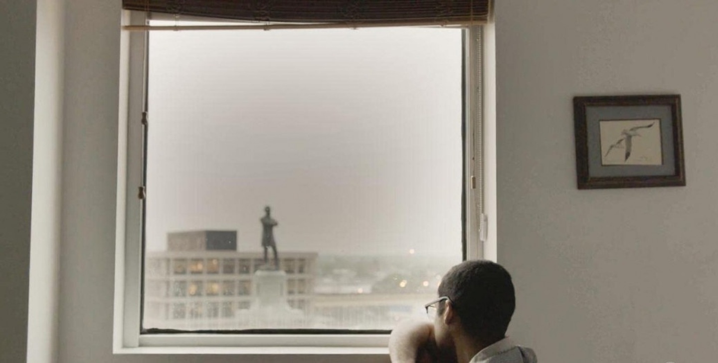 Film still of the head and torso of a person, arm propped against a window, staring outside into a city view, a building and statue prominent in the foreground.