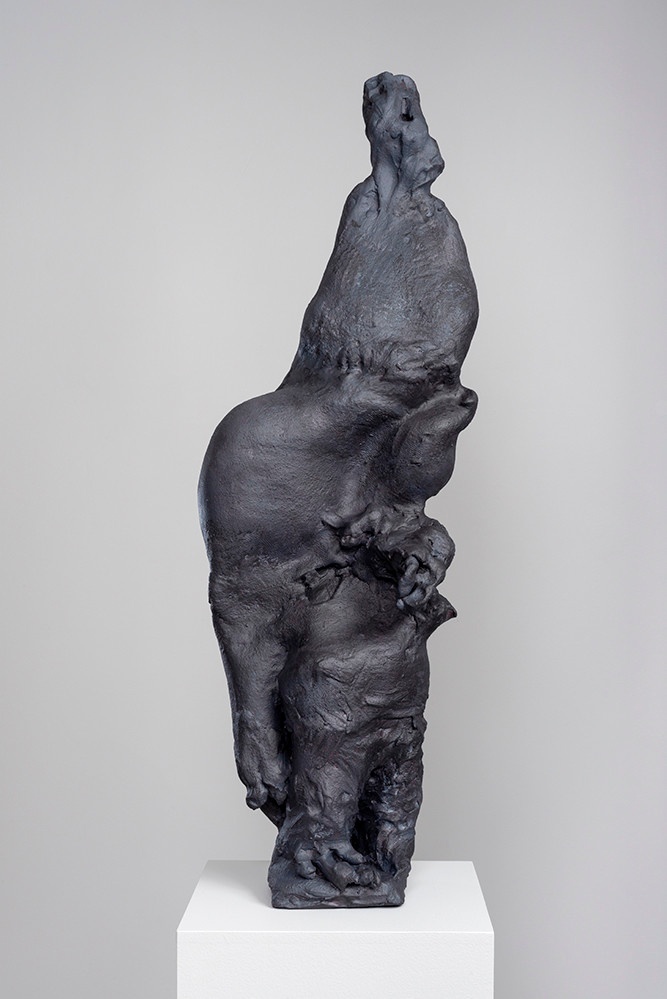 A tall sculpture, in a metallic/gray hue: its form is irregular yet suggestive of human flesh, with intricate folds and masses. It is placed on a white platform.