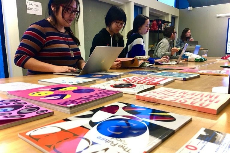 Students at laptops standing at a table with artwork laid across it.