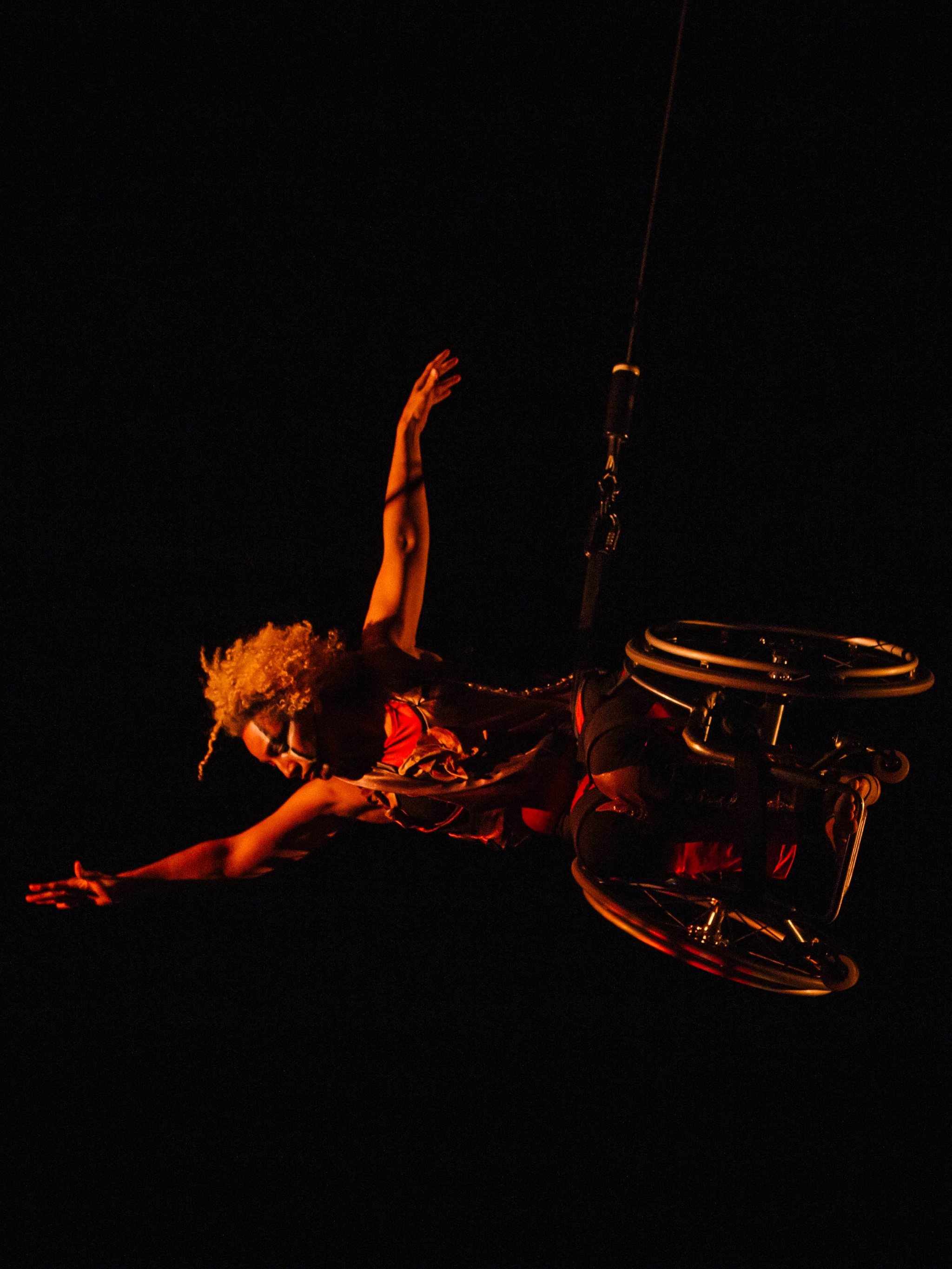 Alice Sheppard flies in her wheelchair, body parallel to the stage as she reaches one arm high and extends the other to the side. She dances in a glowing red shadow. She is a multiracial Black woman with coffee colored skin and short curly hair; she wears a red and gold bodysuit, black and white lines adorn her face.