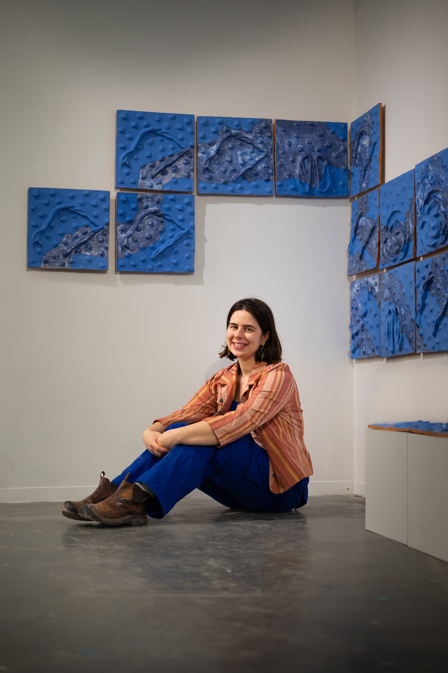 woman with brown shoulder length hair wearing orange sitting on the floor underneath her artwork hanging on the wall