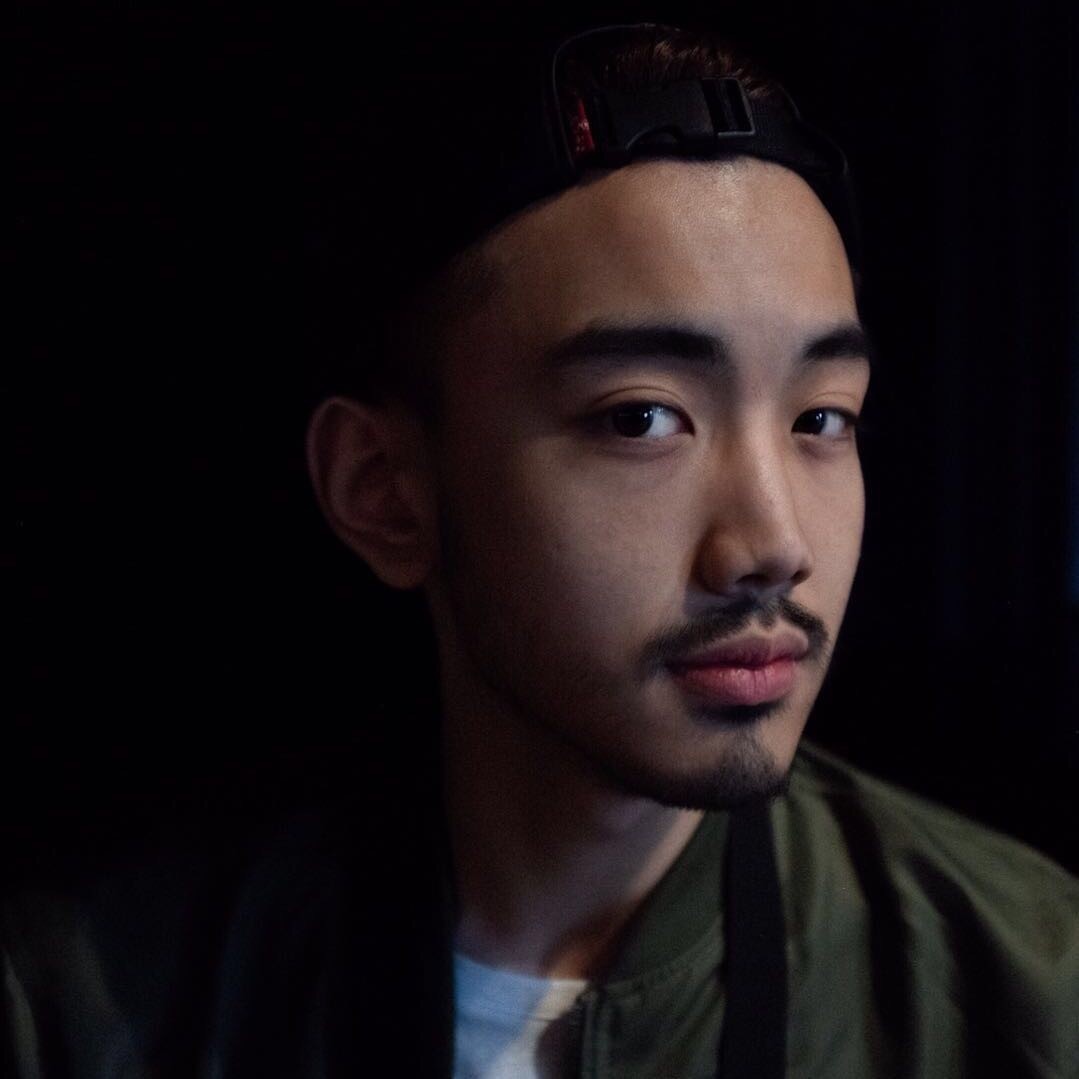A close-up portrait of a young Chinese man seen in three-quarters view. He looks into the camera, wears a cap and green jacket, and has a mustache and goatee. 