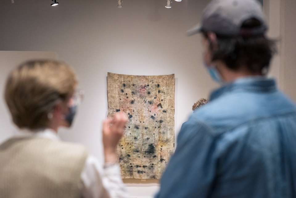 Over the shoulder view of two people looking at a patchwork burlap wall hanging with black spots and stains on it.