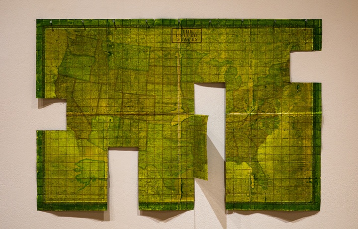 Green-dyed map of the United States that has been stitched with a grid of squares. Several four by four sections of the map have been cut away.