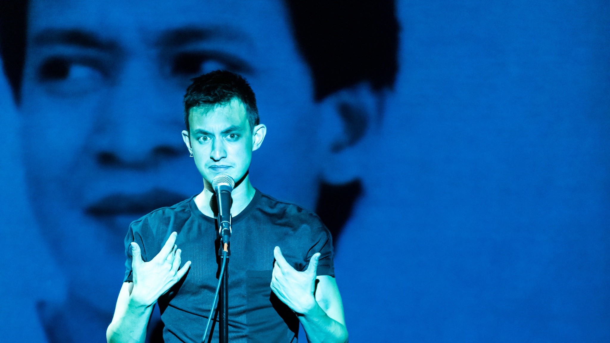 The artist Kyle Dacuyan, a Filipino performer with short dark hair, stands before a microphone bathed in a turquoise light. He trains his gaze intently at us with his mouth drawn tightly closed. He holds his hands up to his chest as if to gesture toward himself. On the wall behind him, an image of a man’s face is projected in a cerulean light at a larger than life scale. 