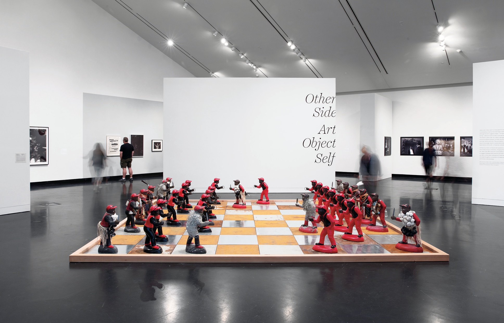 A large metal and wood chessboard with dark-skinned lawn jockeys in black and red clothing acting as chess pieces. Some lawn jockeys are covered in cement with nails, or covered in bottles, or holding knives. 
