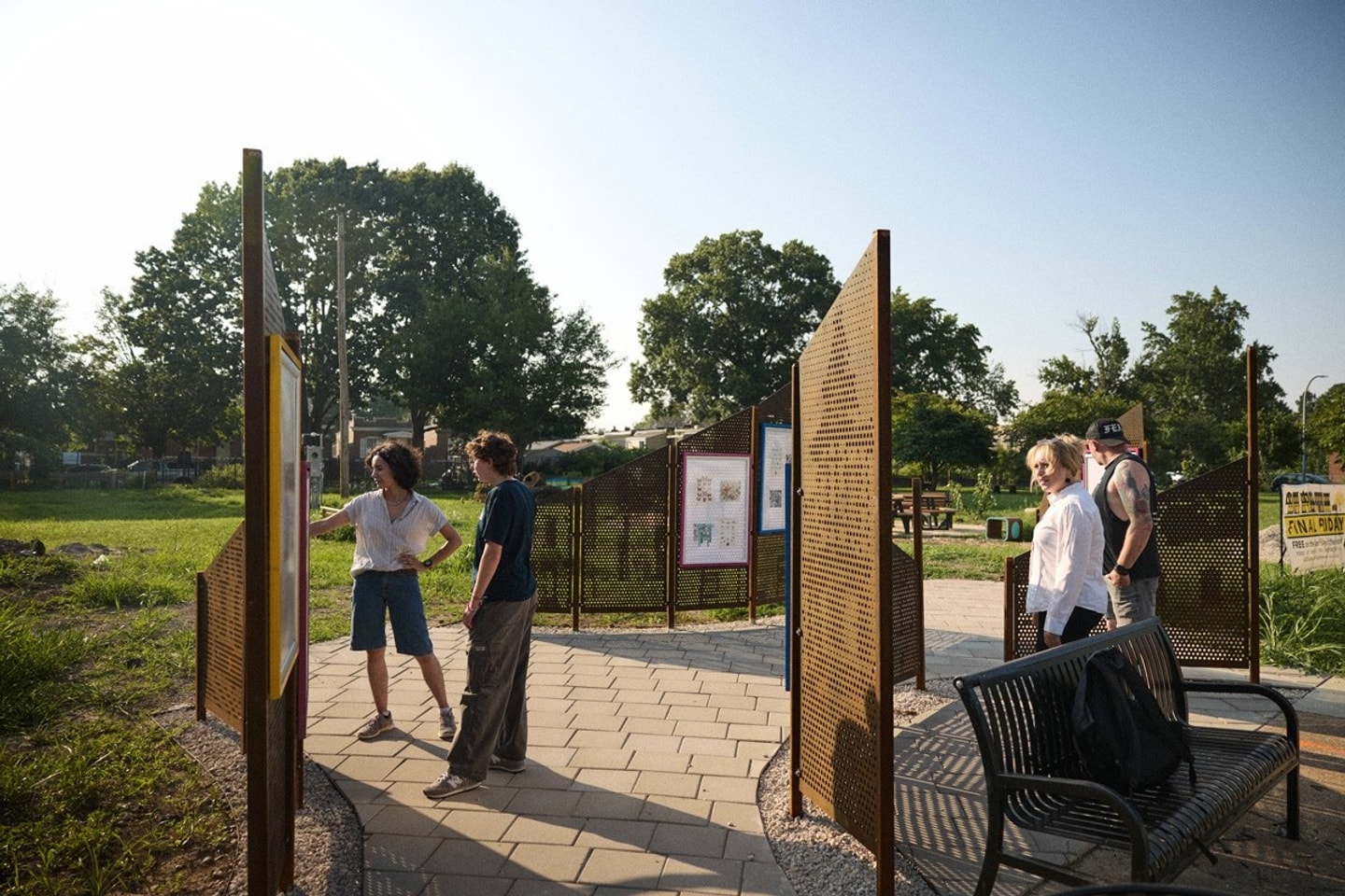 Four people explore an outdoor park installation with paved stone pathways and tall, perforated metal display panels under bright sunlight.