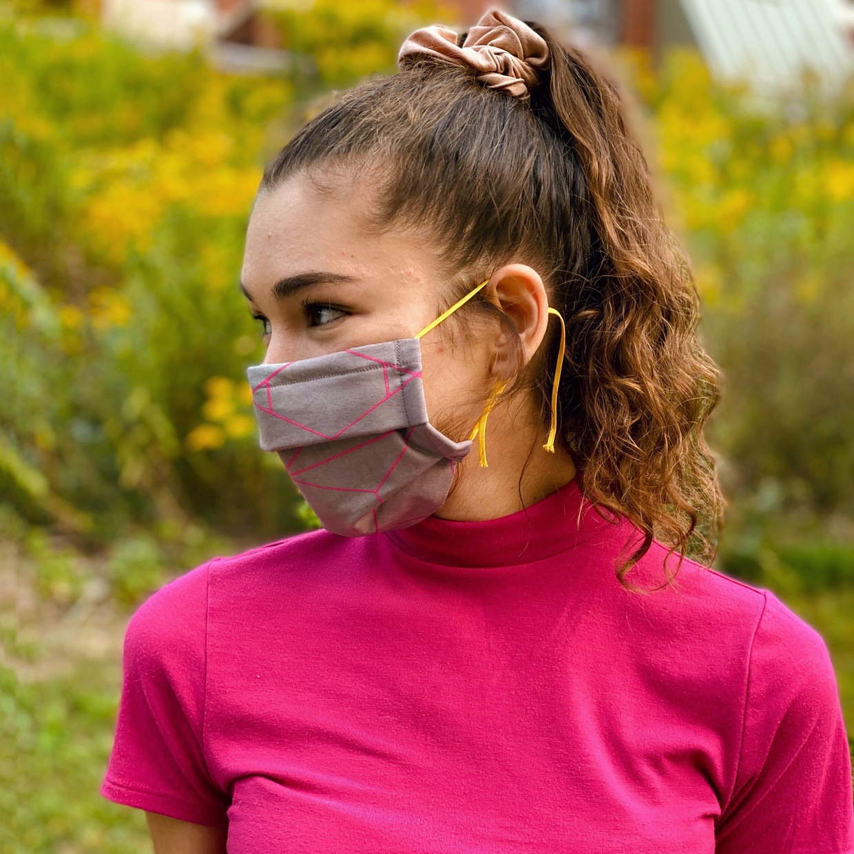 A tanned, young woman with dark hair wears a bright pink shirt and a colorful mask.