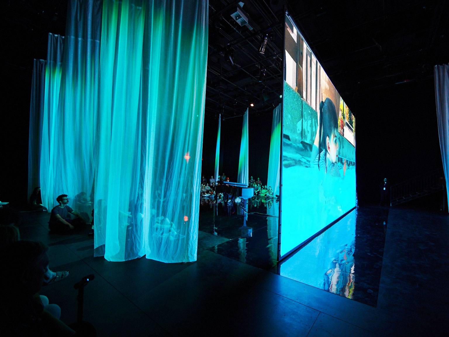 Audience viewing film on large screen in blue-lit space.