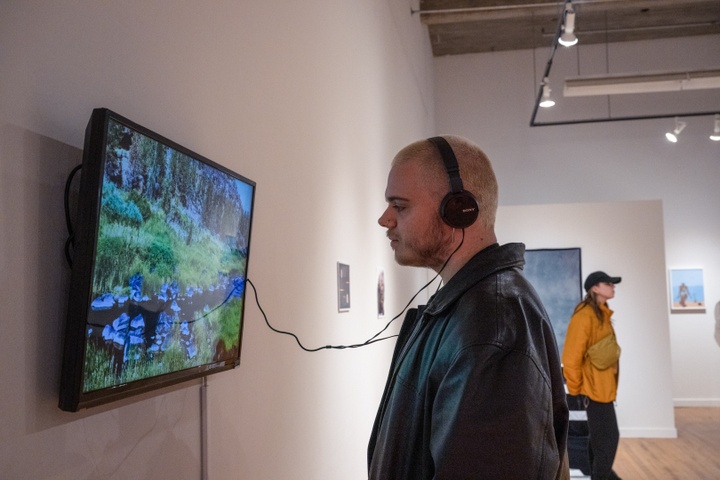 Student wears headphones and stands in front of a wall-mounted video screen displaying an image of a mountain stream.
