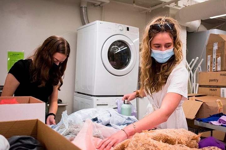 Two students wearing Covid masks sort through piles of fabric remnants on a table. A stacked, white washery/dryer are in the background.