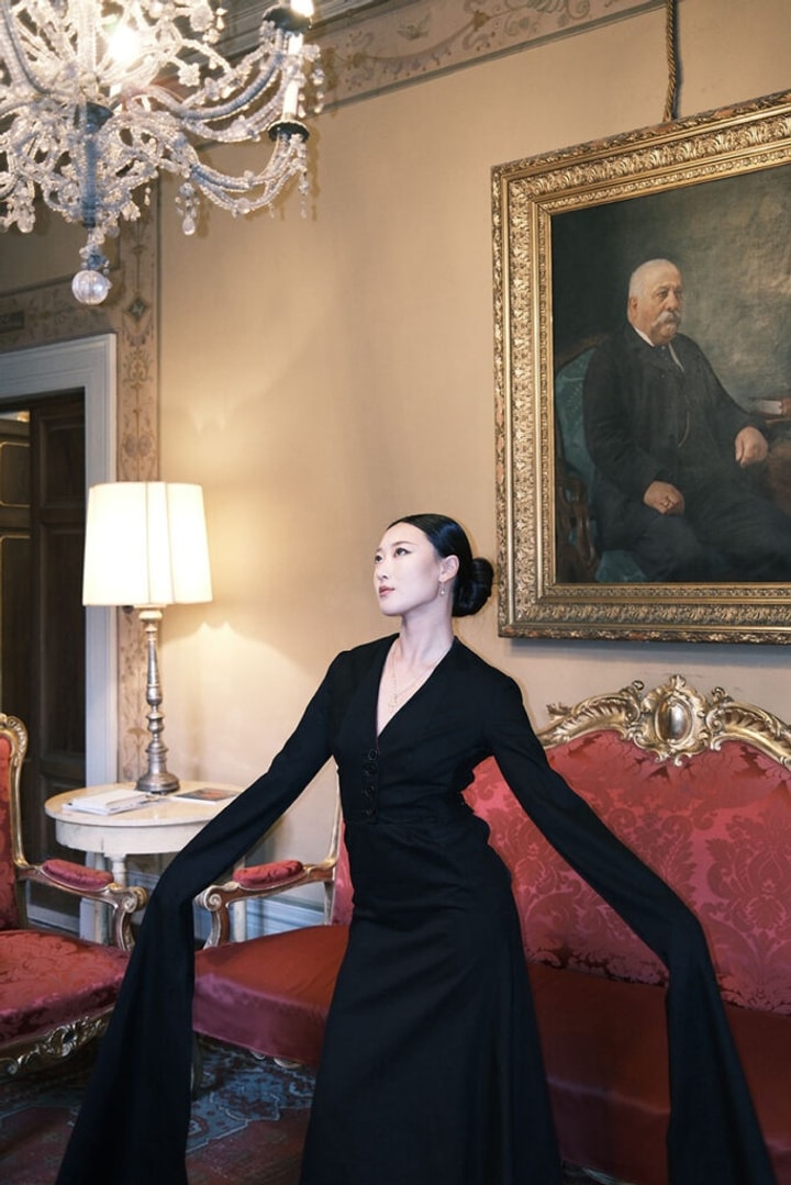 A model poses wearing a long sleeve black dress in a formal room with a crystal chandelier and an oil painting on the wall behin dher.