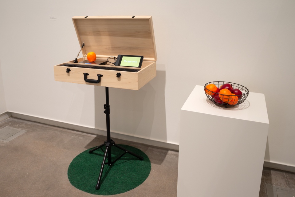 A wooden travel case is displayed on a tripod, opened to reveal a kitchen scale and a tablet. A piece of fruit is on the scale and the tablet displays a text graphic on a yellow background. A basket of fruit sits on a plinth to the right.