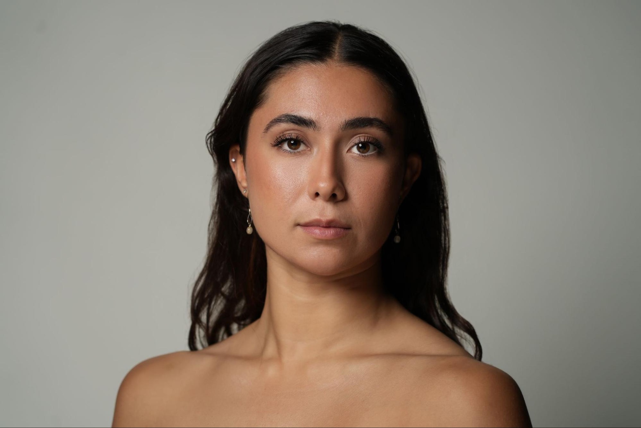 A headshot of dancer Cassandra D’Agostino. She is seen from the shoulders up and has long dark brown hair that falls behind her bare shoulders. She looks directly at us with intention. 