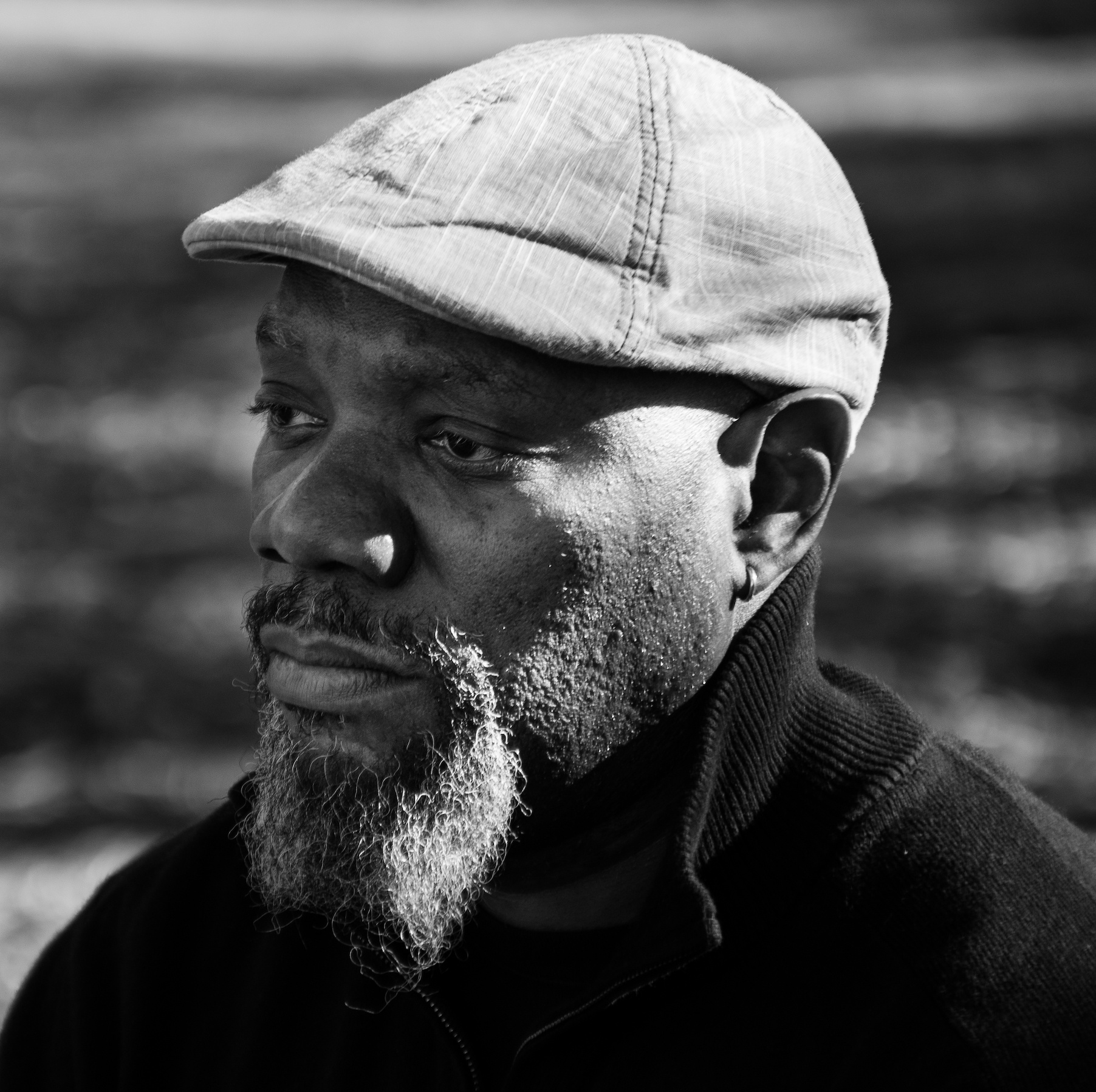 A photo of Mychal Johnson in a high-collared black sweater and cap with sunlight shining from behind him