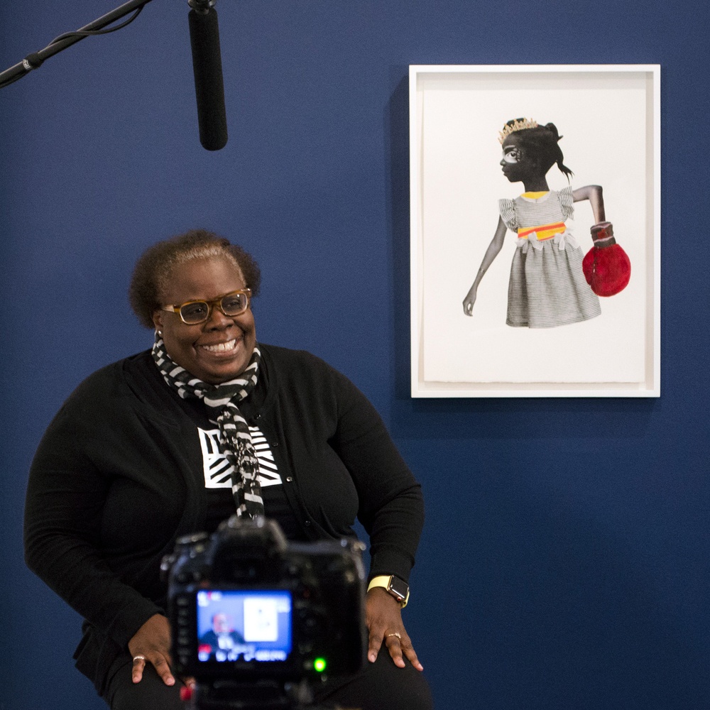 Photograph of Deborah Roberts, a black female artist, laughing in front of a blue wall with her work, Glass Castles, in the background. Glass Castles is an image of a young black girl collaged from various images with a red boxing glove on her left hand.