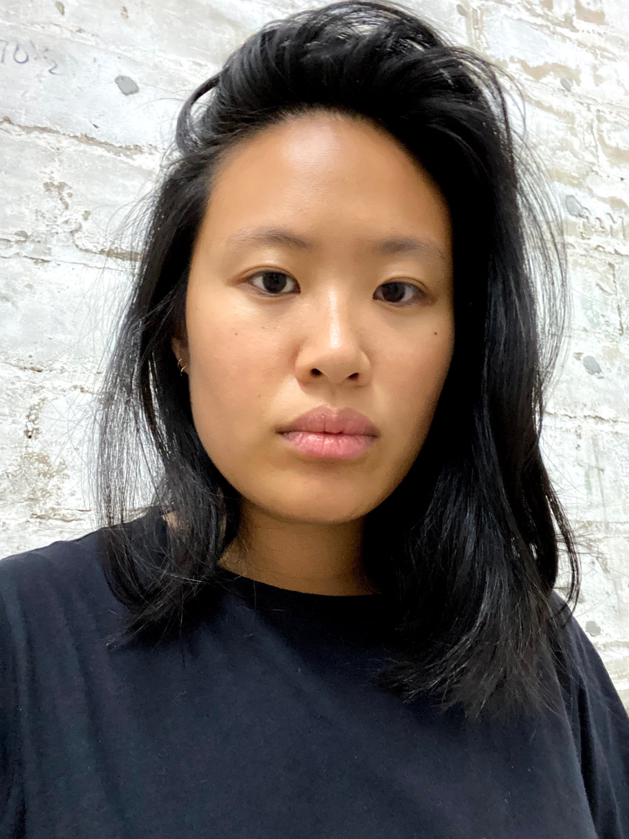 A photo of the artist Pauline Shaw looking into the camera. Shaw's hair is parted to the side and she is in front of a whitewashed brick wall.