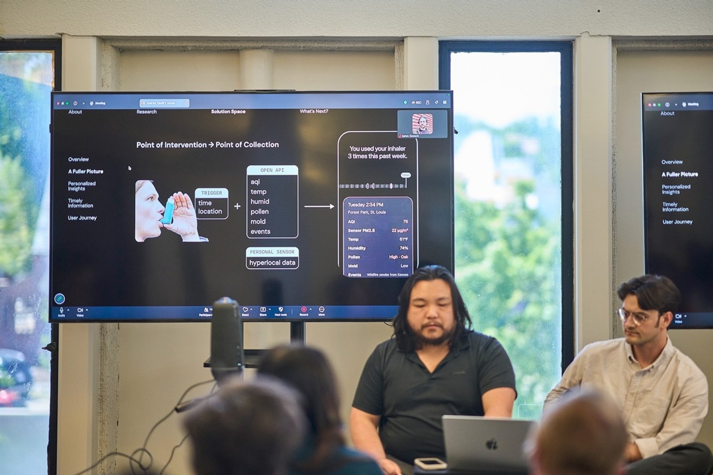 Two males sit in front of a laptop with research projected on a screen behind them and present to a group of people.