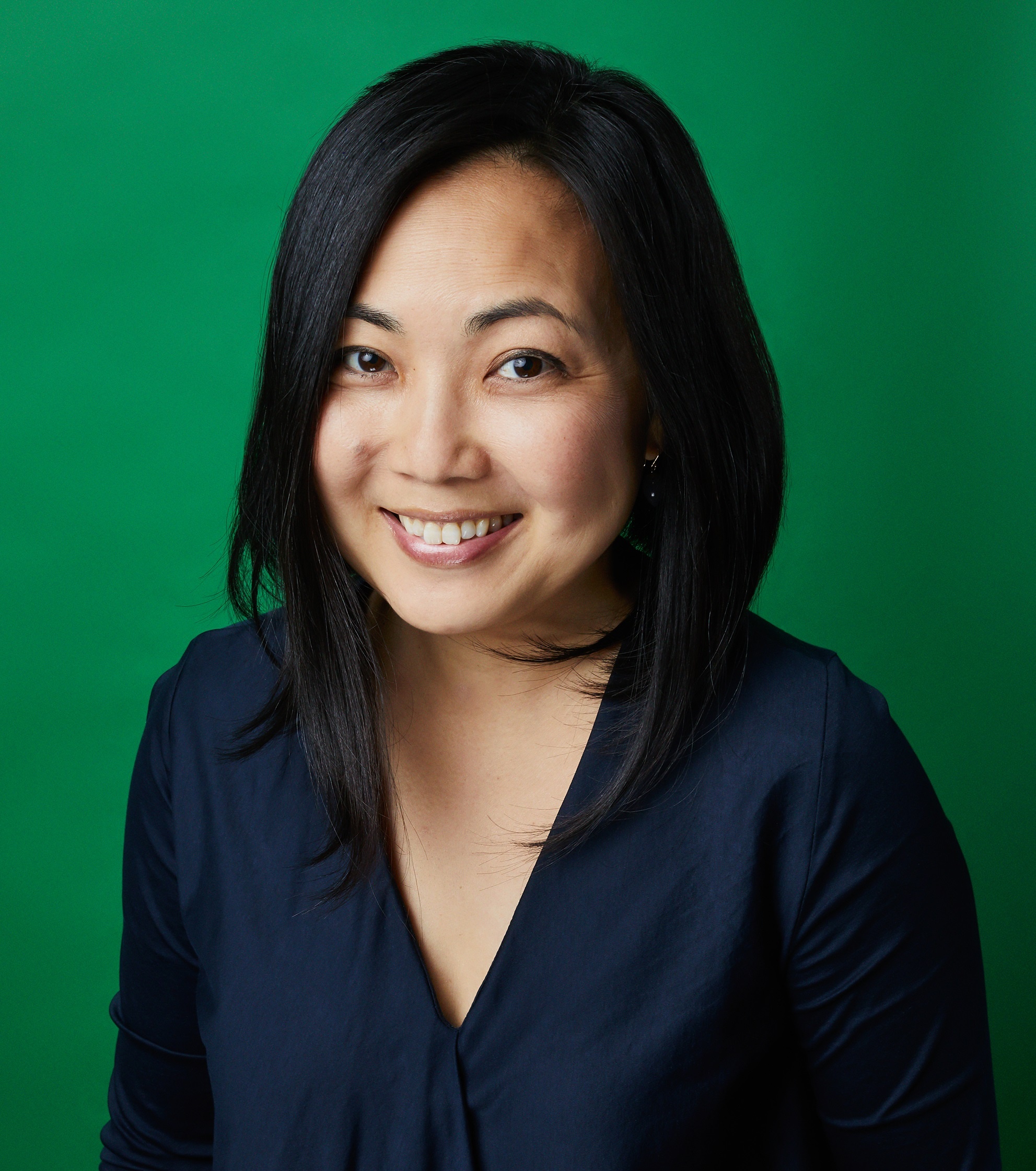 A portrait of Lisa Kim against an emerald background. Lisa has dark hair to her shoulders, wears a navy blue blouse, and smiles at us. 