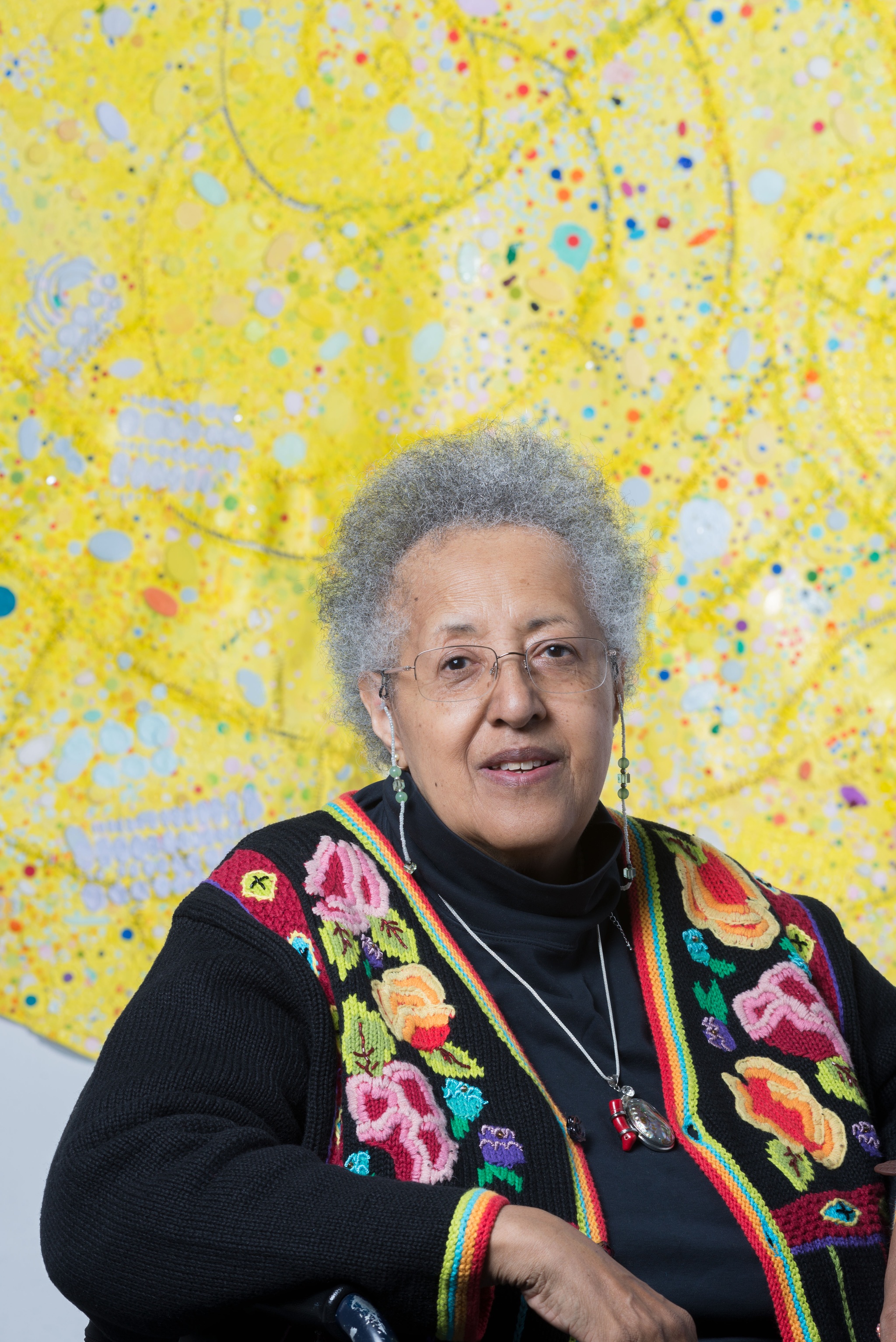 The artist Howardena Pindell sitting in front of one of her painting "Nautilus #1", a curvilinear, yellow, abstract canvas. Pindell wears a colorful cardigan over a dark-colored shirt. 