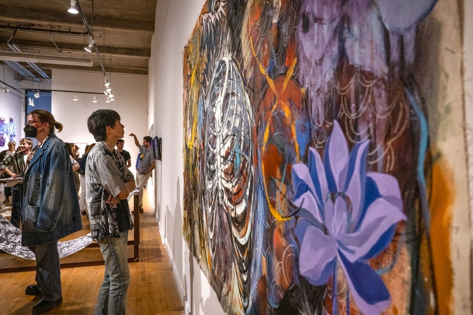 Person stands in a gallery space looking at a large painting on the wall. The painting is colorful with a sketchy, graffiti-like quality. A purple lotus and a skeleton's ribcage are prominent in the design.