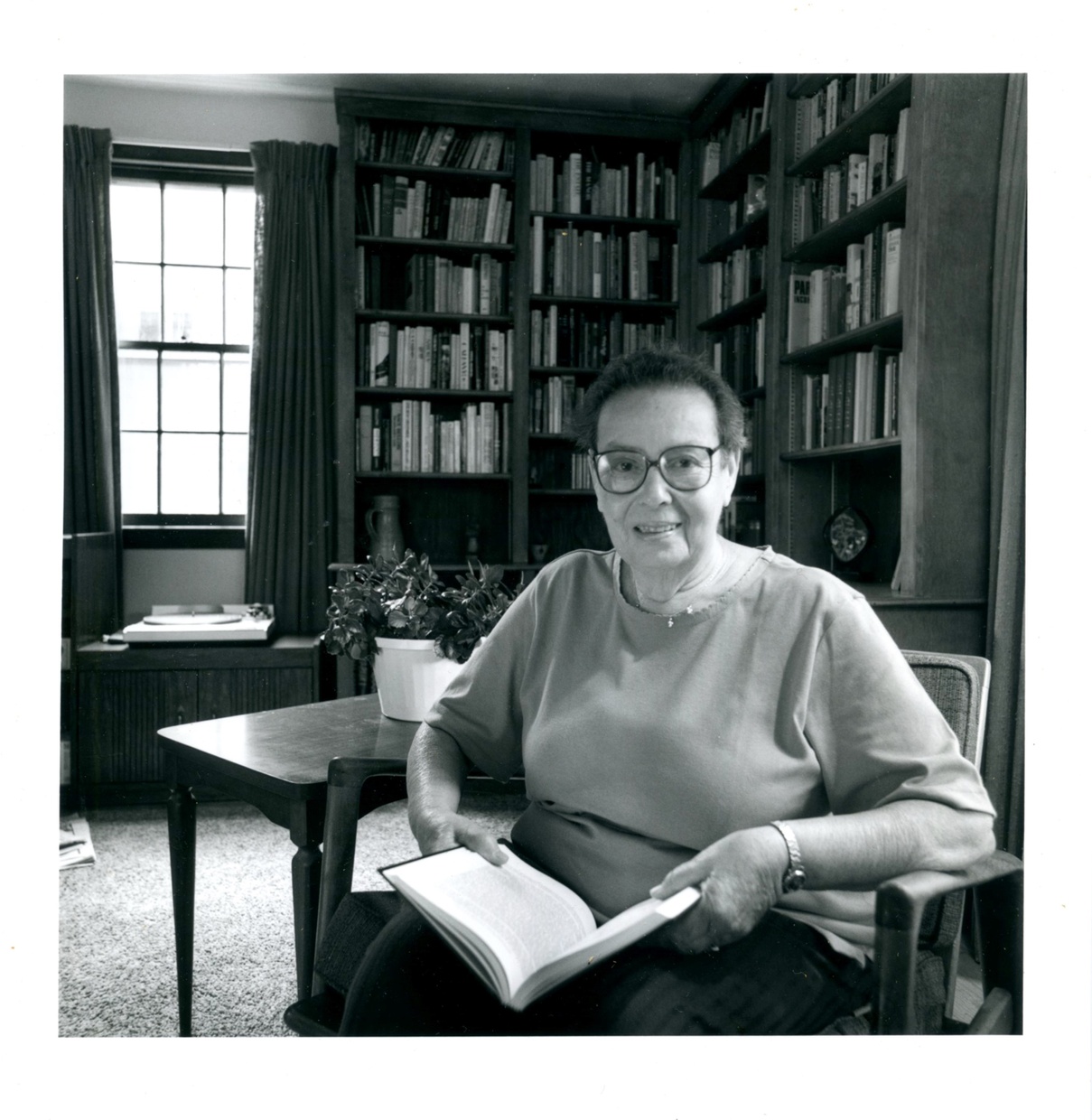 A black and white photograph depicts an older light-skinned woman with glasses smiling and sitting in a chair holding an open book in a room with many bookshelves.