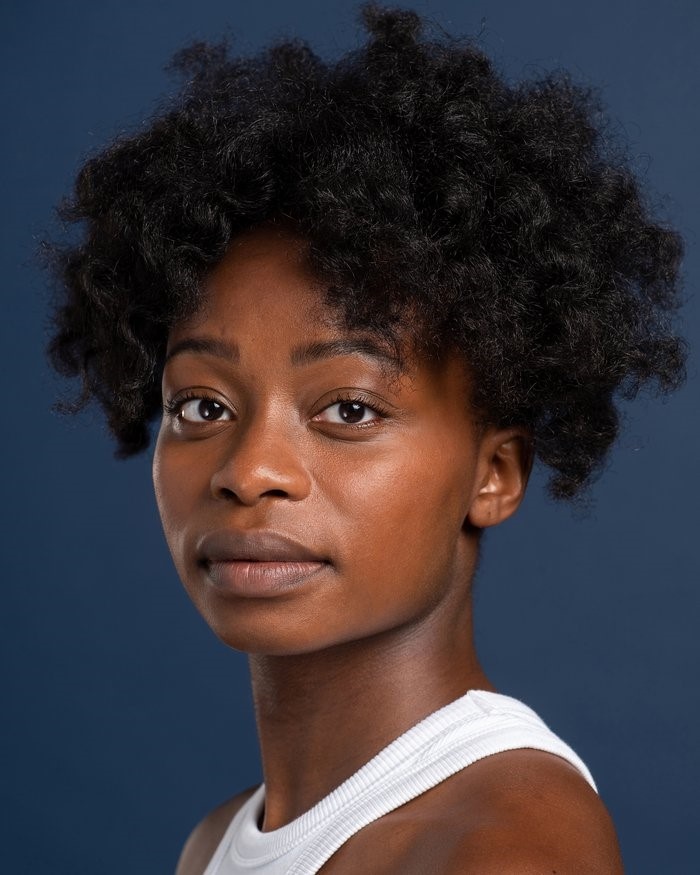 A headshot of actor Deborah Alli, who turns her head to look directly at us. Deborah is Black with dark brown skin and curly black hair. She wears a white tank top. 